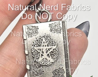 Puede incluir: Un amuleto de libro plateado con un diseño de estrella de cinco puntas y detalles intrincados. El amuleto está unido a una cadena plateada. El texto "Natural Nerd Fabrics Do NOT Copy" es visible en el fondo.