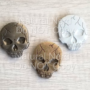 May include: Three skull-shaped decorative objects. One is silver, one is bronze, and the third is white. Each skull has a cracked texture. The skulls are arranged on a light wood-grain surface.