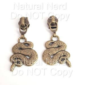 May include: A pair of bronze zipper earrings with snake charms. The snakes are coiled and have a detailed, textured design.