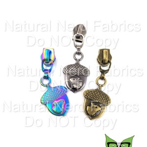 May include: Three zipper pulls with acorn-shaped charms. The charms are silver, gold, and iridescent with a squirrel design. The zipper pulls are for use with clothing or bags.