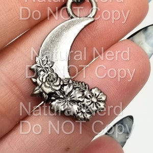 May include: A silver crescent moon charm with a floral design. The charm has a loop at the top for attaching to a necklace or bracelet.