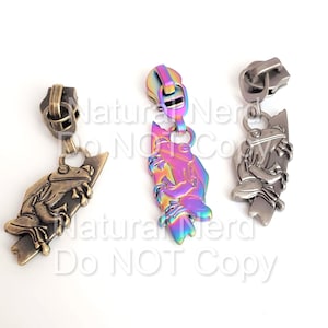 Könnte beinhalten: Drei Reißverschluss-Zipper mit einem Frosch-Design. Der Frosch befindet sich in einer Kletterposition auf einem Ast. Die Reißverschluss-Zipper sind in Antikmessing, Regenbogen-Iridescent und Silber erhältlich.