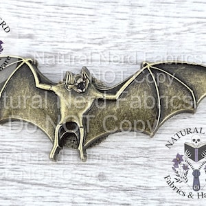 May include: A bronze-colored bat-shaped embellishment with outstretched wings. The bat has detailed features and is likely made of metal. The image includes the text "Natural Nerd Fabrics & Hardware".