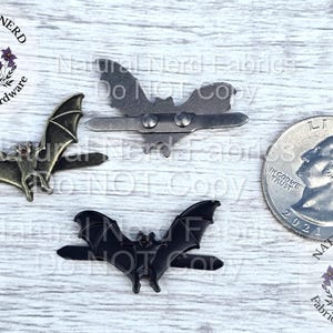 May include: Three metal bat-shaped embellishments in bronze, silver, and black, with a quarter coin for size comparison. The bats have detailed wing designs and are likely for crafting or jewelry making. The image includes the Natural Nerd Fabrics & Hardware logo.
