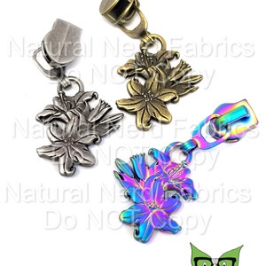 May include: Three zipper pulls with flower designs. The pulls are in silver, gold, and iridescent rainbow colors. The flowers are stylized lilies.