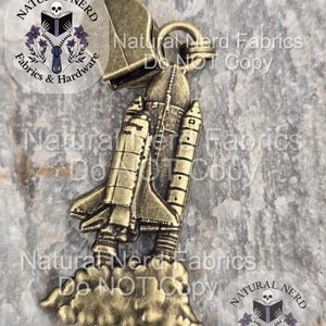 May include: A bronze-colored metal zipper pull featuring a detailed space shuttle design. The shuttle is depicted with wings, engines, and a cloud of smoke. The zipper pull also includes the text "Natural Nerd Fabrics Do NOT Copy".