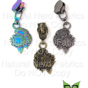 May include: Three zipper pulls with sunflower charms. The zipper pulls are in silver, gold, and iridescent colors. The sunflower charms are detailed with leaves and petals.