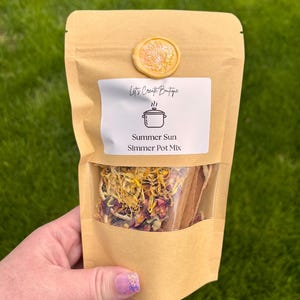 May include: A tan paper pouch containing a Summer Sun Simmer Pot Mix, featuring a clear window displaying dried yellow and pink flower petals and cinnamon sticks. The label includes a pot icon and the text Let's Create Boutique, sealed with a wax seal.