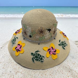 Puede incluir: Un sombrero de paja color canela con ala ancha decorada con apliques de tortugas marinas verdes y flores amarillas con centros rosas. La copa está rodeada por una banda de encaje floral bordado en tonos beige, marrón y verde.