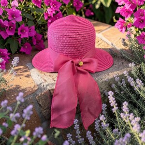Puede incluir: Un sombrero de paja trenzada de color rosa brillante con ala ancha y una cinta de gasa rosa a juego atada en un gran lazo en la parte delantera. Un pequeño dije de metal dorado está sujeto al centro del lazo, añadiendo un detalle metálico.