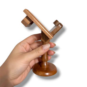 May include: A wooden stand with a round base, a turned stem, and a top section designed to hold an item. The stand is made of light brown wood and has a classic, elegant design. The stand is held by a hand.