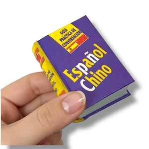 Puede incluir: Un pequeño libro morado titulado "Español Chino" en letras amarillas y blancas. El lomo es amarillo con detalles rojos. El libro también presenta el texto "Guía Práctica de Conversación" en español. El libro se sostiene en una mano sobre un fondo blanco.