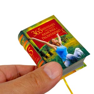 May include: A small, red, yellow, and green book with a yellow ribbon bookmark. The book cover features a woman in a blue tank top and white pants sitting in a field of green grass. The book title is "365 Mensajes para Vivir en Armonia y Prosperidad".