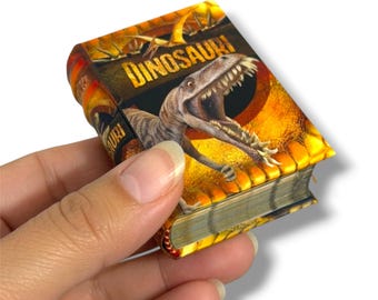 May include: A miniature book titled DINOSAURI featuring a graphic illustration of a dinosaur with an open mouth on the cover. The cover design uses shades of orange, yellow, and brown, with a textured appearance and visible page edges.