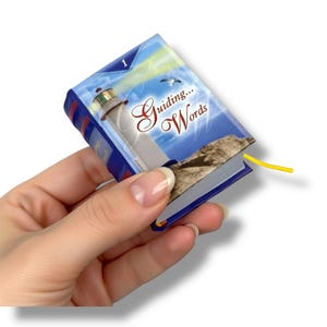 May include: A miniature book titled "Guiding Words" with a lighthouse illustration on the cover. The book has blue and white accents, a yellow ribbon bookmark, and is held in a hand. The cover features the number 1 and a seagull.