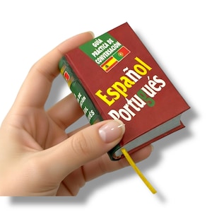 Puede incluir: Un pequeño libro de conversación rojo, titulado "Español Portugués", se sostiene en una mano. El libro tiene una cinta de lectura amarilla y una sección verde con texto y banderas. Está diseñado para hispanohablantes y lusófonos.