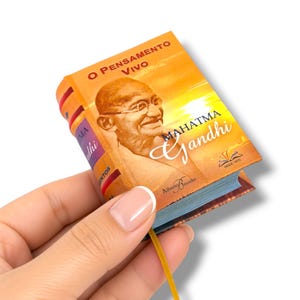 May include: A miniature book titled O Pensamento Vivo Mahatma Gandhi with a portrait of Mahatma Gandhi on the orange and yellow cover. The book features a thin gold ribbon bookmark extending from the bottom and text by Alberto Briceño.