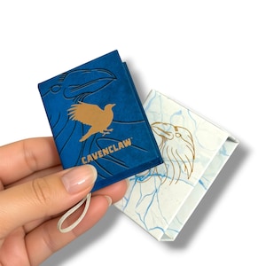 May include: A small blue rectangular box with a gold bird silhouette and the text CAVENCLAW, paired with a white box featuring a blue marble pattern and a gold line-art bird illustration. Both items include a small white fabric loop attached.