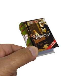 May include: A small, green and red hardcover book titled "Corazón" by Edmundo de Amicis. The book cover features an illustration of a classroom scene with children and a teacher.