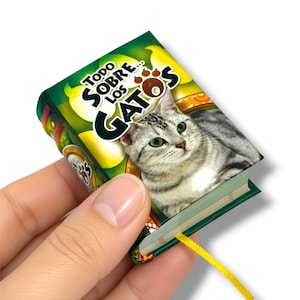 May include: A miniature book titled Todo Sobre Los Gatos featuring a photographic illustration of a grey tabby cat on the cover. The book has a green background, a yellow ribbon bookmark extending from the bottom, and black text on the front cover.