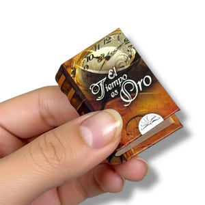 May include: A miniature book with a brown cover and gold-colored spine is held in a hand. The cover features a clock design and the Spanish text "El Tiempo es Oro" in white script. A small white bookmark is visible.