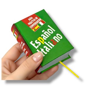 Puede incluir: Un pequeño libro verde titulado "Español Italiano" con una cinta amarilla como marcador. La portada del libro presenta las banderas de España e Italia, junto con el texto "Guía Práctica de Conversación."