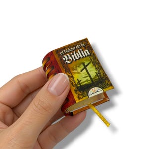 May include: A miniature book titled el Néctar de la Biblia with a brown and gold cover featuring a cross illustration, a decorative border, and a thin yellow ribbon bookmark extending from the bottom edge of the pages.