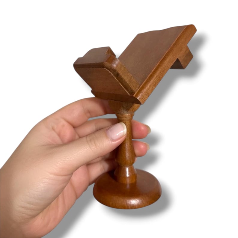 May include: A small, wooden book stand with a dark brown finish. The stand has a round base, a turned stem, and an angled top to hold a book. The stand is held by a hand against a white background.