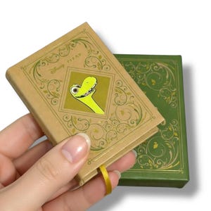 May include: A small, tan-colored book with a gold-colored ribbon bookmark. The cover features a green cartoon character in a gold frame, with the text "Disney Pixar". A second green book is visible in the background.