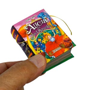 May include: A miniature illustrated book titled "Alicia en el País de las Maravillas" (Alice in Wonderland). The cover features a colorful illustration of Alice in a red dress, with other characters and text. The book has a green spine and a yellow ribbon.