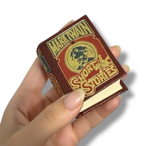 Op de afbeelding: Een miniatuurboek met de titel "MARK TWAIN SHORT STORIES" wordt in een hand gehouden. Het boek heeft een donkerrode kaft met gouden letters en een portret van Mark Twain. De pagina's van het boek zijn lichtcrème van kleur.