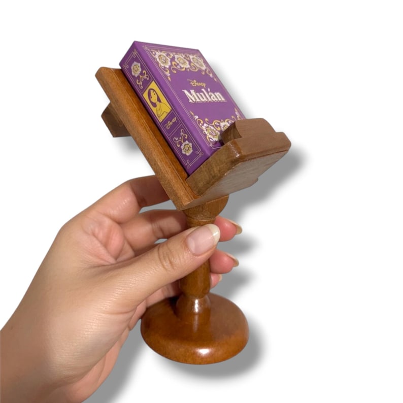 May include: A wooden book stand holding a small purple book with the title "Mulan" in white script. The book stand is made of polished wood and has a round base.