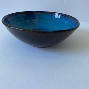 Op de afbeelding: Een kleine, blauwe en zwarte keramische kom met een rode rand. De kom is geglazuurd en heeft een gespikkelde afwerking.