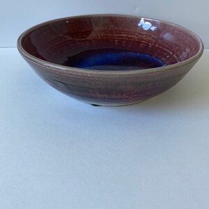 Op de afbeelding: Een grote, ronde keramische kom met een diepe, brede rand. De kom is geglazuurd in tinten van paars, roze en blauw, met een donkerblauw centrum.