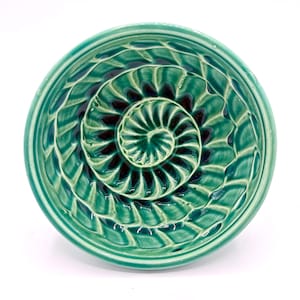 May include: A teal-colored ceramic bowl with a spiral design in the center. The bowl has a textured surface with a repeating pattern of curved lines. The center of the spiral is a darker shade of teal.
