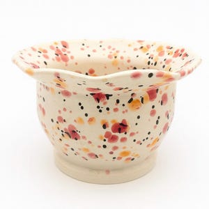 May include: A cream ceramic bowl with a ruffled edge, decorated with a speckled pattern of red, orange, and black dots. The bowl has a rounded base and a flared, wavy edge, creating a decorative and artistic appearance.