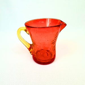 May include: A small glass jug featuring a vibrant red body with a textured, crackled finish and a smooth, translucent yellow handle. The vessel has a wide, open top with a distinct pouring spout and a rounded base, crafted from glass.