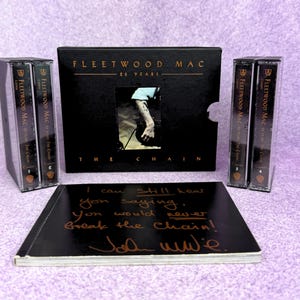 Könnte beinhalten: Ein Fleetwood Mac 25 Years The Chain Box-Set mit vier Musikkassetten in durchsichtigen Kunststoffhüllen, einem schwarzen Schuber mit goldener Schrift und einem zentralen Foto sowie einem schwarzen Booklet mit handschriftlichen Texten und Signatur.