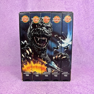 May include: A collection of five black VHS tapes featuring Godzilla movie titles including Godzilla vs. Gigan, Godzilla 1985, Ghidrah The Three-Headed Monster, Godzilla vs. Mechagodzilla, and Son of Godzilla, with graphic illustrations of the monster.