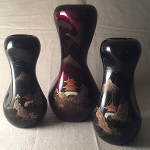 May include: Three black and red ceramic vases with gold painted designs. The vases are all different shapes and sizes. The largest vase is red with gold painted mountains and a pagoda. The two smaller vases are black with gold painted mountains and a pagoda.