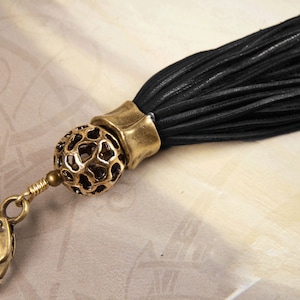 May include: A black leather tassel with a gold metal clasp and a decorative ball charm. The ball charm has a geometric design.