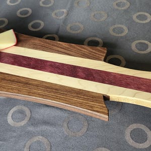 Peut inclure: Une planche de service en bois rectangulaire composée de bandes alternées de noyer brun foncé, d'érable crème clair et de bois pourpre. La planche possède une poignée avec une découpe circulaire et porte un petit bol d'olives, du fromage et des tomates.