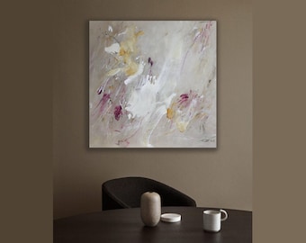May include: Abstract painting with a white background and splashes of pink, purple, and gold paint. The painting is hanging on a wall in a room with a brown chair and a table.