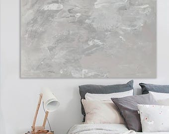 May include: Abstract canvas art in shades of gray and white, mounted above a bed with gray and white pillows and a white comforter. A wooden desk lamp and a stack of books sit on a side table.