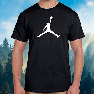 Peut inclure: T-shirt noir avec une silhouette blanche d'un joueur de basket-ball en train de sauter pour attraper un ballon. Le motif est centré sur la poitrine. Le t-shirt est à manches courtes et semble être en coton.