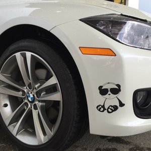 May include: A white BMW car with a black and white panda sticker on the front bumper. The panda is wearing sunglasses. The car has a silver alloy wheel and a black tire.