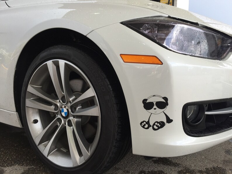 May include: A white BMW car with a black and white panda sticker on the front bumper. The panda is wearing sunglasses. The car has a silver alloy wheel and a black tire.