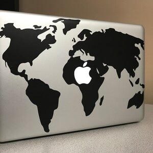 May include: A silver laptop with a black world map design. The Apple logo is visible in the center of the map. The laptop is on a light gray surface.