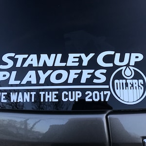 May include: White sticker on a car window with the text "STANLEY CUP PLAYOFFS" above "WE WANT THE CUP 2017". The Oilers logo is on the right side. The sticker is on a dark background.