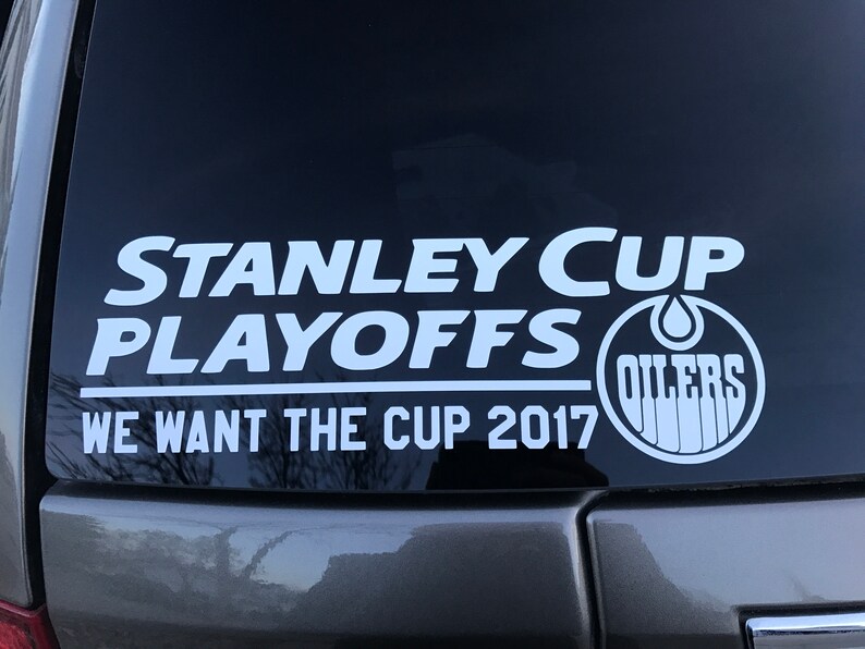 May include: White sticker on a car window with the text "STANLEY CUP PLAYOFFS" above "WE WANT THE CUP 2017". The Oilers logo is on the right side. The sticker is on a dark background.